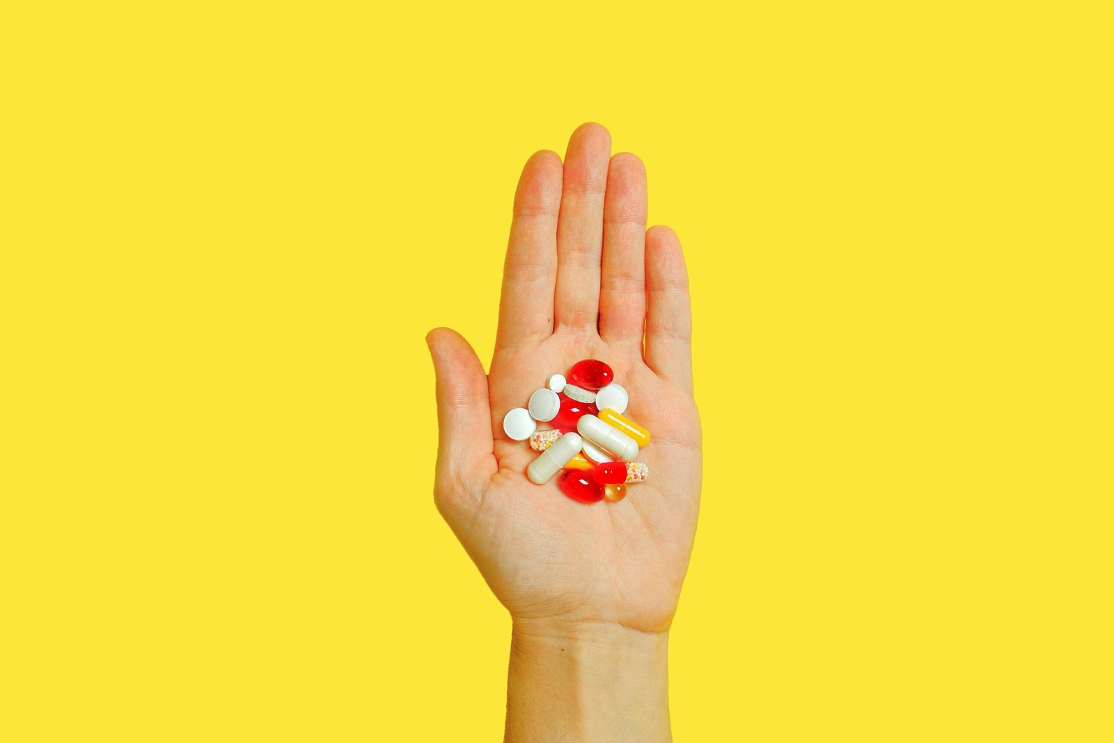 Close-up of an adult hand holding different types of pills against a vivid yellow background.